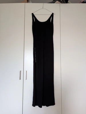 Svart ärmlös jumpsuit med vida ben - Säljer en svart, lång och ärmlös jumpsuit med breda axelband och vida ben. Jumpsuiten har en enkel och stilren design, perfekt för dig som gillar minimalistiskt mode. Materialet ser mjukt och följsamt ut, vilket ger en skön känsla.