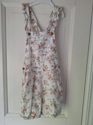 Vit blommig jumpsuit med knappar - Superfin vit jumpsuit med blommigt mönster i rosa och blått. Från Newbie i stl 128. Axelbanden har volangdetaljer och jumpsuiten har träknappar på sidorna samt en bröstficka. Perfekt för dig som vill ha en söt och avslappnad look. Lite schabbig på vissa ställen (syns på sista bilderna), men inget som syns på håll! Ändast använd fåtal gånger!