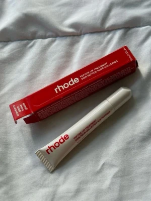 rhode Peptide Lip Treatment - Peptide Lip Treatment från rhode i en vit tub med röd text. Kommer i en matchande röd förpackning. Läppbalsamet har en smidig applikator och är perfekt för att återfukta och vårda läpparna. Tuben är gjord av plast och har en modern, minimalistisk design.