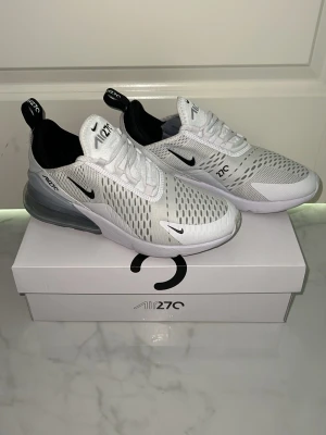 Nike Air Max 270 vita sneakers - Nike Air Max 270 sneakers i vitt med svarta detaljer och stor synlig Air-enhet i hälen. Ovandelen är i mesh för bra ventilation och har en modern, sportig look. Swoosh-logga på sidan och AIR270-tryck på plösen och hälen. Perfekta för dig som gillar streetwear.