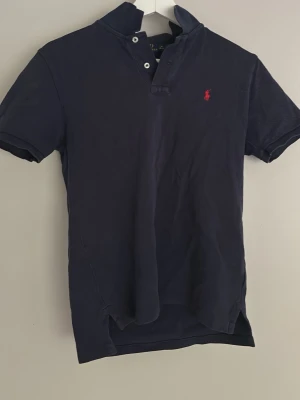Marinblå pikétröja från Ralph Lauren - Klassisk marinblå pikétröja från Ralph Lauren med korta ärmar och röd broderad logga på bröstet. Tröjan har krage, knappar framtill och är tillverkad i bomull för en bekväm känsla. Perfekt till jeans eller shorts.