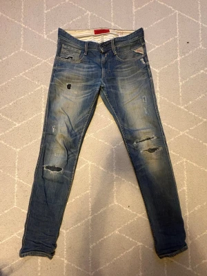 Sjukt snygga replay anbass - Säljer ett par blå jeans med slitningar och hål på benen. Jeansen har en klassisk femficksmodell och tydliga slitningar för en trendig, avslappnad look. Materialet är denim med stretch för extra komfort.