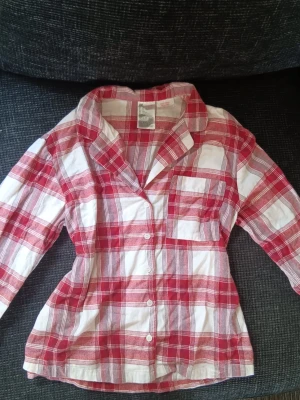 pyjamas set - Klassisk tvådelad pyjamas med långärmad skjorta och byxor i rödvitt rutigt mönster. Skjortan har krage, bröstficka och knappar framtill. Byxorna har dragsko i midjan för extra komfort. Perfekt till jul och mysiga kvällar.
