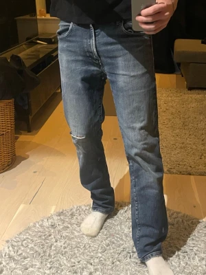 Blå raka jeans med slitning - Säljer ett par blå jeans med rak passform och lätt slitning på ena benet. Jeansen har klassisk femficksdesign och normal midja. Perfekta för en avslappnad och trendig look.
