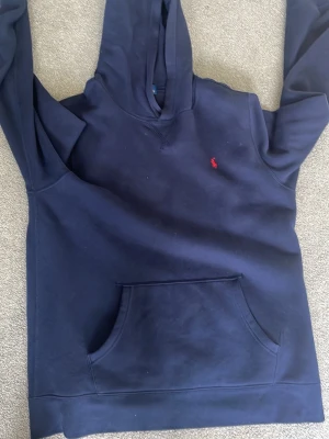 Mörkblå hoodie från Polo Ralph Lauren - Snygg mörkblå hoodie från Polo Ralph Lauren med klassisk röd logga broderad på bröstet. Hoodien har en stor känguruficka framtill och en skön huva. Tillverkad i mjukt bomullsmaterial som passar perfekt för chill dagar. Storlek XL för yngre, passar dig i längd 170