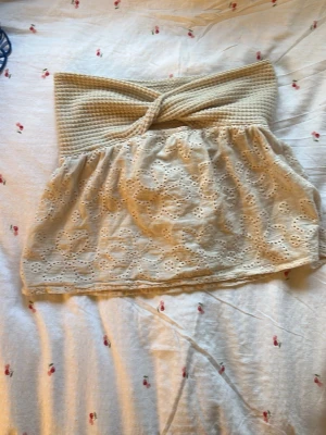 Beige spets tubtopp med twist - Söt beige tubtopp med twistad överdel i ribbad struktur och nedre del i broderad spets med blommönster. Perfekt för sommardagar och enkel att styla med jeans eller kjol.
