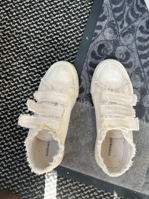 Beiga sneakers med fransade kanter - Säljer ett par beiga sneakers med fransade kanter och tre kardborreband. Skorna har en tjock vit sula och är tillverkade i tygmaterial. Perfekta för en avslappnad och trendig stil. Använder knappt och har ej orkat tvättat och därför säljer jag för billigare. Använt ungefär två gånger!💗