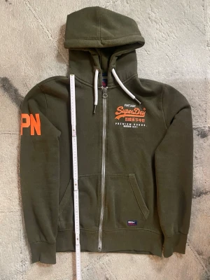 ”Unik” Grön Superdry Zip Hoodie - Olivgrön hoodie från Superdry med vit dragkedja, huva med vita snören och orange tryck på bröstet samt ärmen. Mjuk insida och ficka framtill. OBS alla bilder noggrant. 