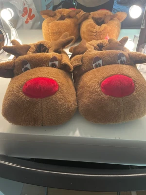Rudolf med röda mullen tofflor - Mysiga tofflor i mjuk plysch med renmotiv (Rudolf med röda mullen). Tofflorna är bruna med stora röda nosar, horn och broderade ögon. Perfekta för att ge en lekfull touch till hemmastilen, speciellt under vintern och julen. Passar mig me sl 39 men passar även mindre fötter