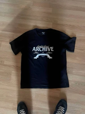 Svart Jack & Jones ARCHIVE t-shirt - Snygg svart t-shirt från Jack & Jones Essentials med ARCHIVE-tryck i vitt framtill. Klassisk rund hals och korta ärmar. Perfekt till jeans eller shorts för en clean och enkel stil.