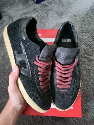 Svarta sneakers från Diesel med röda snören - Unika svarta sneakers från Diesel med grov textur på ovandelen och snygga röda snören. Skorna har beige sula och detaljer i svart skinn samt Diesel-logga på sidan och plösen. Perfekta för dig som vill sticka ut med din stil.