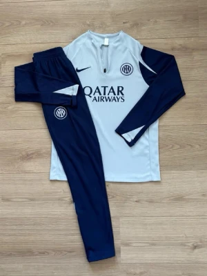 Nike Inter träningsset blå/vit - Snyggt träningsset från Nike med Inter-logga. Tröjan är ljusgrå med mörkblå ärmar och dragkedja vid halsen. Byxorna är mörkblå med vit detalj och Inter-emblem. Materialet är lätt och andas, perfekt för fotbollsträning eller gym. S 160-172 cm ish