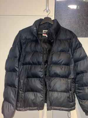 Svart dunjacka med Thermore-foder - Snygg svart dunjacka med quiltad design och hög krage. Jackan har dragkedja framtill, två sidofickor med knapp och innerfoder av Thermore Ecodown Fibers som håller värmen. Perfekt för kalla vinterdagar och har en stilren look.