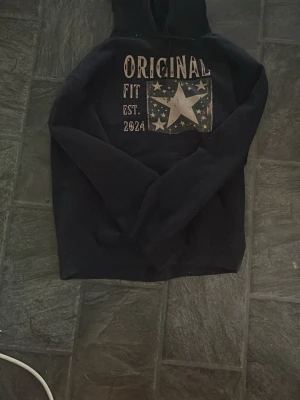 Marinblå hoodie från original archive  - Marinblå  hoodie med huva och stort tryck på bröstet där det står 'ORIGINAL FIT EST. 2024' samt en stor vit stjärna och mindre stjärnor i bakgrunden. Klassisk passform och långa ärmar. Perfekt för en avslappnad stil.