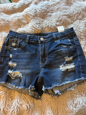 Mörkblå slitna jeansshorts - Säljer ett par mörkblå jeansshorts med slitna detaljer och fransig kant. Shortsen har klassisk femficksdesign, knapp och dragkedja framtill samt kontrastsömmar. Perfekta för sommaren och ger en avslappnad vibe.