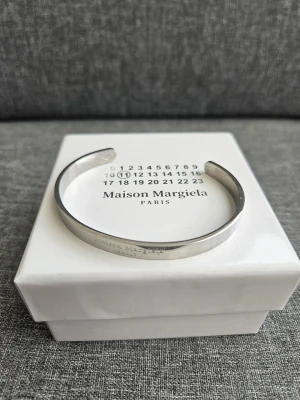 Maison Margiela silverfärgat armband - Snyggt och minimalistiskt armband från Maison Margiela i silverfärgad metall. Öppen design som gör det enkelt att ta på och av. Diskret logotyp ingraverad på utsidan och märkning på insidan. Perfekt accessoar för en clean och modern stil.