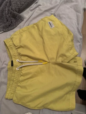 Gula shorts med snörning - Säljer ett par ljusgula shorts med elastisk midja och vit snörning med gula detaljer. Shortsen har en avslappnad passform och är perfekta för varma dagar. Tillverkade i ett mjukt bomullsmaterial som känns skönt mot huden.
