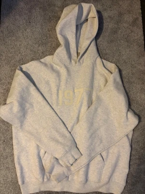EssentialsHoodie - Snygg ljusgrå hoodie med trycket '1977' på bröstet. Hoodien har en stor huva, ribbade muddar och en klassisk känguruficka framtill. Perfekt för en avslappnad och trendig stil.