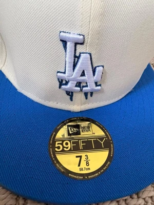 Vit och blå New Era LA keps - Snygg 59FIFTY keps från New Era med LA-logga framtill. Kepsen är vit med blått broderi och har en blå skärm samt knapp på toppen. Materialet är klassisk bomullscanvas och sidan har broderad text i blått.