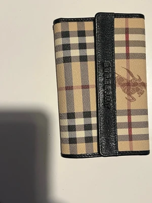 Burberry plånbok i klassiskt rutmönster - Snygg plånbok från Burberry i beige, svart och rött klassiskt rutmönster med svarta läderdetaljer. Plånboken har präglad Burberry-logga på framsidan och är tillverkad i en kombination av läder och canvas. Perfekt accessoar för dig som gillar stilrena detaljer.