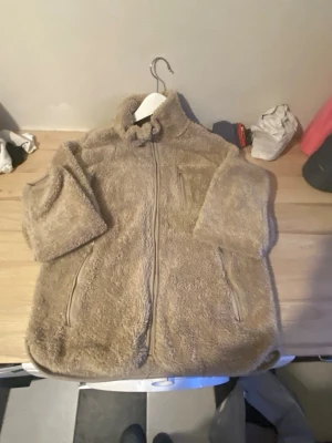 Beige teddyjacka från Marine XS - Mysig beige teddyjacka från Marine i storlek XS (32/34). Jackan har dragkedja framtill, två sidofickor med dragkedja och hög krage. Tillverkad i fluffigt teddy-material som håller dig varm och bekväm. Perfekt för kyliga dagar och enkel att matcha med olika outfits. Har dock ett litet håll i vänstra ficka.