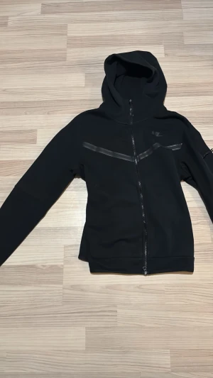 Svart hoodie med dragkedja - Säljer en svart hoodie med hel dragkedja och huva. Hoodien har diskreta detaljer i form av svarta linjer över bröstet och är tillverkad i ett mjukt material. Perfekt för en avslappnad och stilren look.