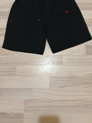 Svarta shorts med röd logga - Snygga svarta shorts med en liten röd logga broderad på vänster sida. Klassisk passform och enkel design, perfekta för sommaren eller träning. Mjukt material och elastisk midja för extra komfort.