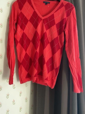 Röd argyle stickad tröja Tommy Hilfiger - Snygg röd stickad tröja från Tommy Hilfiger med klassiskt argyle-mönster i olika röda nyanser och tunna vita linjer. Tröjan har V-ringning och långa ärmar, perfekt för en trendig och avslappnad look.