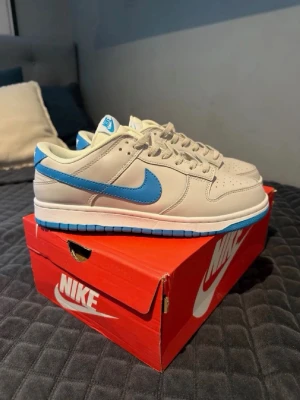 Nike Dunk Low med blå swoosh - Nike Dunk Low sneakers i beige med blå swoosh och detaljer. Skorna har en klassisk låg siluett, perforerad tå och vit mellansula med blå yttersula. Tillverkade i läder med textildetaljer och plös med Nike-logga.