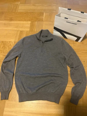 Grå halvzip-tröja från J.Lindeberg - Snygg grå stickad tröja från J.Lindeberg med halv dragkedja och hög krage. Tillverkad i mjukt material som ger en clean och stilren look. Perfekt att bära över en t-shirt eller skjorta för en avslappnad men ändå uppklädd stil.