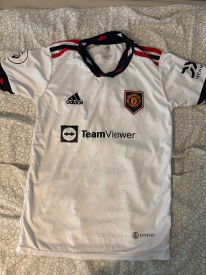 Manchester United tröja  - Snygg vit Manchester United bortatröja från Adidas med svarta och röda detaljer. Tröjan har klubbmärke, sponsortryck och Ronaldo 7 på ryggen. Tillverkad i lätt och ventilerande material, perfekt för fotboll eller träning.