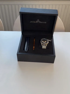 Jacques Lemans  klocka - Säljer en helt ny Jacques Lemans Chronograph herrklocka som aldrig har varit använd eller buren. Klockan kommer i originalförpackningen och extra läderarmband samt verktyg för att byta armband ingår.  * Modell: Jacques Lemans Chronograph * Skick: Helt ny, aldrig använd * Originalbox ingår * Silverfärgat mesharmband * Extra läderarmband ingår * Verktyg för armbandsbyte ingår * Mörk urtavla med silverfärgade och gula detaljer * Passar både till vardags och finare tillfällen