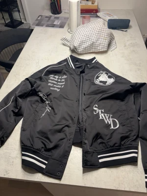 Svart bomberjacka Pull&Bear M - Svart bomberjacka från Pull&Bear med vita tryck och broderier framtill. Jackan har vita ränder på muddar och krage, samt coola detaljer och text på bröstet. Tillverkad i glansigt syntetmaterial och har klassisk bomberjacke-look.