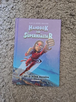 Handbok för superhjältar Del 2 - Handbok för superhjältar Del 2: Röda Masken av Elias & Agnes Våhlund. Boken har ett färgstarkt omslag med en tecknad superhjälte i röd dräkt mot en blå och lila bakgrund. Inbunden bok från Rabén & Sjögren, perfekt för dig som gillar spännande äventyr.