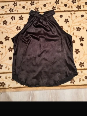 Svart halterneck topp i satin - Snygg svart halterneck topp i glansigt satinliknande material. Toppen är ärmlös och har hög krage med knäppning i nacken. Perfekt för dig som vill ha en stilren och trendig look.