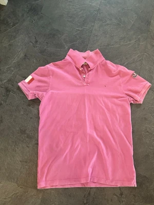 Moncler piké Rosa - Snygg rosa pikétröja från Moncler med korta ärmar och klassisk krage. Moncler-logga på ärmen och broderad text på kragen. Perfekt för dig som vill sticka ut med en färgglad och trendig topp till sommaren. Små fläkar men troligen tvättbara med vilja! 
