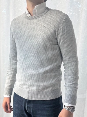 Ljusgrå stickad tröja - Snygg ljusgrå stickad tröja med diskret mönster och rund halsringning. Tröjan har ribbade muddar vid ärmslut och nederkant, samt en liten broderad logga på bröstet. Perfekt att bära över en skjorta för en clean och stilren look. Storleken är L men funkar även för storlek M. 