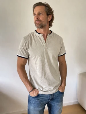 Beige kortärmad t-shirt från Zara - Snygg beige t-shirt från Zara med korta ärmar och kontrasterande mörkblå kant vid hals och ärmslut. T-shirten har knappar vid halsen för en extra detalj och är tillverkad i mjuk bomull. Perfekt för en avslappnad och stilren look.