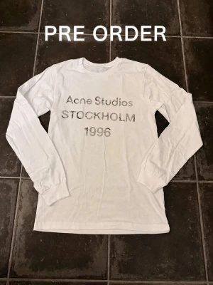 Vit långärmad Acne Studios t-shirt - Vit långärmad t-shirt från Acne Studios med tryck i grått framtill där det står 'Acne Studios STOCKHOLM 1996'. Klassisk passform och rund halsringning. Perfekt för dig som gillar minimalistisk och stilren design. Kommer runt 6e september. Är ca 182 och väger 67
