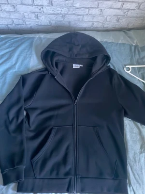 Svart zip hoodie från Andy by Frank Dandy - Säljer en svart hoodie med dragkedja från Andy by Frank Dandy. Hoodien har huva, två stora fickor framtill och ribbade muddar vid ärmslut och nederkant. Tillverkad i mjukt material som känns skönt mot huden. Perfekt för en chill och avslappnad stil.