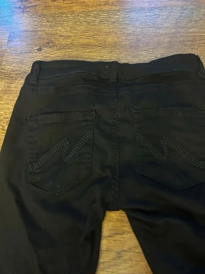 Svarta bootcut jeans med detaljer - Säljer ett par svarta bootcut jeans med snygga nitar i zigzag-mönster på bakfickorna. Klassisk femficksmodell med knapp och dragkedja framtill. Jeansen har breda ben och är tillverkade i ett stretchigt material för skön passform. Använd en gång