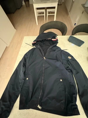 Mörkblå vindjacka Moncler - Snygg mörkblå vindjacka med huva och dragkedja framtill. Jackan har två fickor med knappstängning och en diskret logga på ärmen. Materialet är lätt och passar perfekt för blåsiga dagar. Stilsäker detalj med randig kant på huvan.
