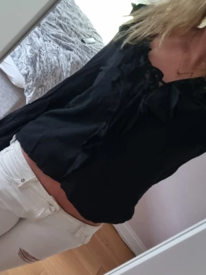 Svart blus med knyt och volanger - Snygg svart blus med långa ärmar, volangdetaljer och knytning vid halsen. Blusen har en luftig passform och är tillverkad i ett lätt tyg som ger en avslappnad look. 