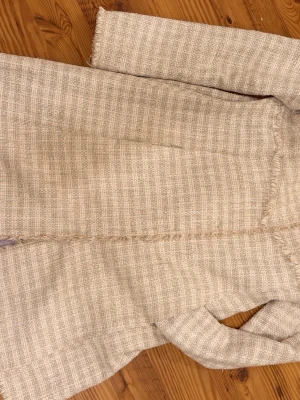 Beige tweedkappa med fransar från Odd Molly - Stilren retro beige kappa i tweed med fransade kanter och diskreta knappar framtill. Kappan har ett ljust lila innerfoder och en snygg passform. Perfekt för dig som gillar klassisk men trendig stil. En fläck på framsidan som inte syns när kappan är stängd. Se bild Storleken är 3 hos Odd Molly 