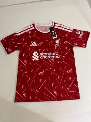 Liverpool röd matchtröja Adidas - Snygg röd Liverpool matchtröja från Adidas med vitt mönster och vita detaljer på axlarna. Tröjan har klubbmärke och sponsortryck framtill samt korta ärmar. Tillverkad i lätt och ventilerande material, perfekt för träning eller match.