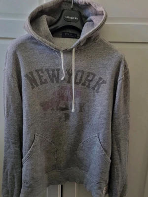 Polo Ralph lauren vintage hoodie - Snygg grå hoodie från Polo Ralph Lauren med huva och dragsnören. Stor New York-tryck framtill och känguruficka. Mjuk bomullsmix som känns skön mot huden. Detaljerad etikett på ärmen för extra stilpoäng.