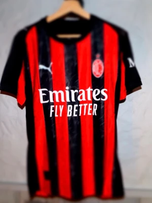 AC Milan matchtröja röd/svart Puma - Snygg AC Milan matchtröja från Puma i klassiska röda och svarta vertikala ränder. Tröjan har klubbmärke på bröstet, Puma-logga och Emirates Fly Better-tryck på framsidan. Tillverkad i lätt och ventilerande material, perfekt för fotboll eller träning. Är du efter en annan storlek är det bara att skriva!