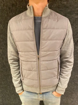 Grå reiss cardigan med dragkedja - Snygg grå cardiganjacka med quiltad front och ryggparti i mjukt sweatshirtmaterial. Jackan har ståkrage, ribbade muddar och två fickor med dragkedja. Perfekt för lager på lager och en clean streetstil.