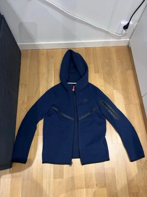Mörkblå Nike tech - Snygg mörkblå hoodie med hel dragkedja framtill och huva. Jackan har två snedställda fickor med dragkedja på bröstet och en extra ficka med dragkedja på ena ärmen. Perfekt för en sportig och avslappnad stil.