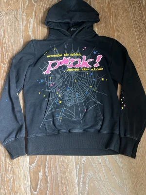 Svart hoodie med spindelnätsprint - Cool svart hoodie med huva och färgglad print framtill. Texten 'nevermind the spider, pink! here’s the slime' och ett stort spindelnät i vitt, dekorerad med stjärnor i rosa, blått och gult. Mjuk bomullskänsla och känguruficka framtill. Perfekt för dig som gillar edgy streetwear.